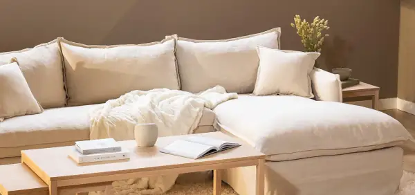 En bekväm soffa med pastell inredning, prydd med mjuka kuddar och en fluffig pläd. Ett soffbord i trä med böcker och en vas fulländar den lugna atmosfären.