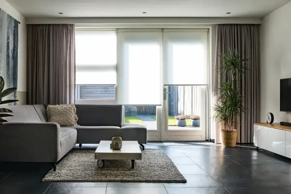 Grå soffa i vardagsrummet med draperade gardiner och stora fönster som släpper in ljus.